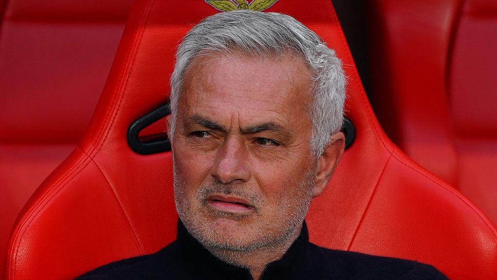 Close-up of a grey-haired man with stubble sitting in a red stadium seat, wearing a black top, with a wary expression.