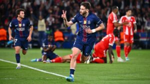 Paris Saint-Germain player in blue jersey celebrates a goal, flashing a peace sign as teammates and opponents look on from the field's edge.