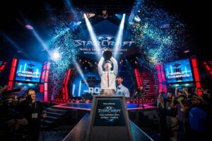 Esports player in a white jacket raises a trophy on a brightly lit stage with blue confetti during the StarCraft World Championship Series finals, crowd cheering in the foreground.