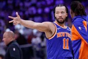 Basketball player in a blue Knicks jersey (#11) extending his arm and pointing during a game on the court and crowd in the background.