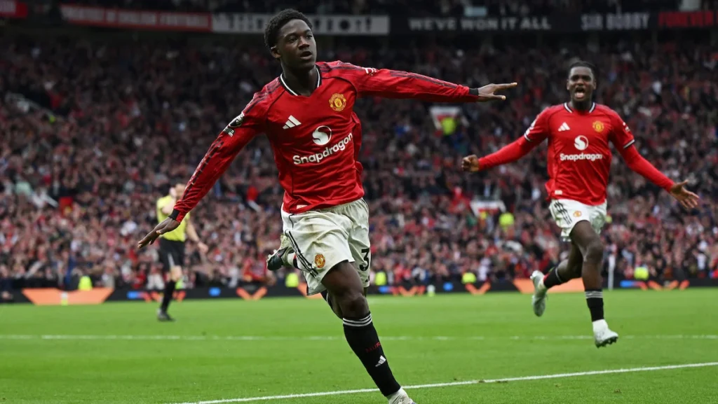 Two Manchester United players celebrate a goal on a packed stadium, foreground player mid-run with arms outstretched.