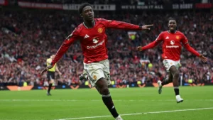 Two Manchester United players celebrate a goal on a packed stadium, foreground player mid-run with arms outstretched.