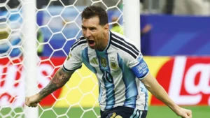 Male soccer player in an Argentina-style blue-and-white striped jersey celebrates a goal near the net.