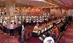 Busy casino scene with rows of slot machines and players seated or standing at them in a colorful, carpeted hall.