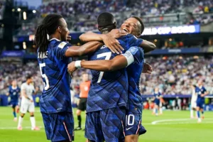 Three soccer players in blue jerseys hug and celebrate on the field after a goal, crowd in the stands behind them (numbers 5, 7, and 10 visible).