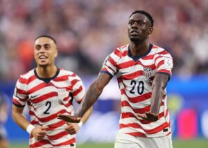 Two USA soccer players in red-and-white striped jerseys celebrate on the field.