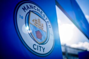 Close-up of the Manchester City crest on a blue banner, showing the ship and red rose emblem.