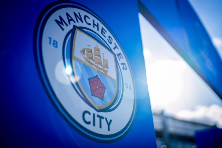 Close-up of the Manchester City crest on a blue banner, showing the ship and red rose emblem.
