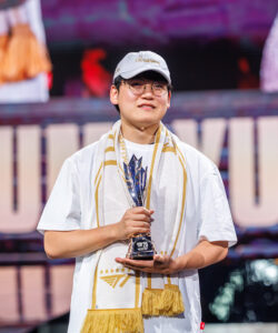 Person on stage wearing a white shirt and cap, holding a tall trophy with a gold scarf draped over the shoulders, smiling at the camera.